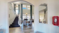 This impressive home showcases an open, airy design anchored by a dramatic two-story great room with soaring ceilings, floor-to-ceiling plaid drapery panels, and an expansive bank of windows that flood the space with natural light. A sweeping staircase with white balusters and a dark carpet runner serves as a focal architectural centerpiece, leading to an upper-level overlook. The living area is thoughtfully arranged with comfortable seating, wood-look flooring, and wall sconces that lend a warm ambiance. A stone-clad fireplace with a dark wood mantel adds texture and character to one corner of the main living space, accompanied by a glass-paned French door that opens to the outdoors. The kitchen is sleek and modern, featuring dark cabinetry, quartz countertops, a mosaic tile backsplash, and a full suite of stainless steel appliances. A dedicated home theater room delivers an immersive entertainment experience, complete with rows of plush leather reclining seats, classic movie poster artwork, a bold red accent wall, and a playful bean bag chair. The cohesive use of light wood flooring, neutral tones, and carefully chosen furnishings throughout creates a refined yet welcoming atmosphere suited for both relaxation and entertaining.