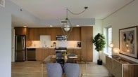 This thoughtfully designed single-floor residence blends contemporary aesthetics with warm, curated details throughout. The kitchen is a standout space, showcasing rich wood cabinetry that extends to the ceiling for ample storage, paired with a classic white subway tile backsplash and stainless steel appliances. A geometric brass pendant light anchors the room, casting a warm glow over the glass-topped dining table with its gold base and modern upholstered chairs. A potted fiddle-leaf fig tree adds a natural, organic element to the space. The light wood-tone flooring ties the open layout together seamlessly. The bedroom offers a striking contrast, featuring deep charcoal accent walls that create a moody, sophisticated atmosphere. A tufted, channel-stitched upholstered headboard serves as the focal point, flanked by windows that frame views of lush palm trees and fill the room with natural light. Above, a faceted geometric pendant light with a frosted glass finish adds an artistic touch. Curated artwork adorns the walls, while layered bedding in soft teal, white, and blush tones completes the refined, design-forward look of the room.