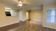 This single-floor residence offers a clean, functional layout designed for comfortable everyday living. The main living area is anchored by rich, dark hardwood-style flooring that runs throughout, providing a warm contrast against the soft, neutral beige walls and crisp white trim and baseboards. Crown molding adds a refined architectural detail to the space, while a ceiling fan with a light fixture ensures both comfort and practical illumination.

The open layout allows for a seamless transition between the living space and the adjacent kitchen, which is partially visible through a pass-through counter with a granite-style surface — a standout feature that adds both utility and visual interest. The kitchen area is accessible via a nearby doorway, contributing to the overall flow of the floor plan.

Track lighting in the secondary area provides targeted illumination, complementing the overall lighting design of the space. Doorways lead to additional rooms, suggesting a well-connected and easily navigable layout. The neutral color palette throughout creates a bright, versatile backdrop that adapts well to a variety of interior styles. Overall, the space presents a tidy, move-in-ready atmosphere with thoughtful finishes and a cohesive design aesthetic.