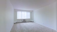 This single-floor space presents a clean, minimalist interior centered around a spacious living area. The room is finished with wall-to-wall neutral carpeting that extends throughout, contributing to a soft, quiet ambiance underfoot. Crisp white walls and a smooth white ceiling create a bright, airy atmosphere that feels open and uncluttered.

A large picture window serves as the focal point of the main living area, fitted with vertical blinds that allow flexible control of natural light. Below the window sits a wall-mounted HVAC unit flanked by baseboard heating units, providing both heating and cooling directly within the room. This self-contained climate control setup ensures year-round comfort.

The layout of the room is open and versatile, offering ample space for a variety of furniture arrangements. The absence of architectural obstructions within the main area allows for flexible use of the space. Overall, the interior maintains a neutral, functional aesthetic with practical features integrated throughout, making it a straightforward and adaptable living environment.