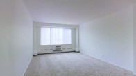 This single-floor space presents a clean, minimalist interior centered around a spacious living area. The room is finished with wall-to-wall neutral carpeting that extends throughout, contributing to a soft, quiet ambiance underfoot. Crisp white walls and a smooth white ceiling create a bright, airy atmosphere that feels open and uncluttered.

A large picture window serves as the focal point of the main living area, fitted with vertical blinds that allow flexible control of natural light. Below the window sits a wall-mounted HVAC unit flanked by baseboard heating units, providing both heating and cooling directly within the room. This self-contained climate control setup ensures year-round comfort.

The layout of the room is open and versatile, offering ample space for a variety of furniture arrangements. The absence of architectural obstructions within the main area allows for flexible use of the space. Overall, the interior maintains a neutral, functional aesthetic with practical features integrated throughout, making it a straightforward and adaptable living environment.