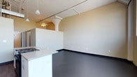 This single-floor space showcases an open-concept loft design that blends industrial and modern aesthetics. The layout is characterized by its generous ceiling height, which adds a sense of airiness and volume throughout the unit. Exposed ductwork and track lighting run along the ceiling, reinforcing the industrial character of the space, while a pendant light fixture adds a softer touch overhead.

The dark, polished concrete flooring anchors the space with a sleek, contemporary feel and contrasts cleanly against the bright white walls. A partial wall with a rounded column creates a natural division between the living area and the kitchen zone, adding architectural interest without fully enclosing the space.

The kitchen area features a countertop peninsula with a built-in cooktop, offering a functional workspace that opens toward the main living area. The white cabinetry and light countertop surfaces complement the minimalist design palette. A ceiling fan is visible in an adjacent zone, providing both comfort and additional lighting. The overall ambiance is modern and understated, with a neutral color scheme that provides a versatile canvas for a variety of uses.