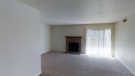 This single-floor space centers around a living area that combines classic and functional design elements. The room is anchored by a traditional brick fireplace with a wooden mantel, serving as a natural focal point and adding warmth and character to the space. Adjacent to the fireplace, a large sliding glass door fitted with vertical blinds allows for generous natural light to fill the room, while also providing access to the outdoors. The carpeted flooring in a neutral gray tone runs throughout, contributing to a soft, comfortable underfoot feel. Walls are finished in a clean off-white, complementing the understated color palette of the space. The textured popcorn ceiling adds a layer of depth overhead, consistent with the architectural style of the unit. The open layout of the living area offers flexibility for various furniture arrangements. Overall, the space presents a straightforward and functional design, well-suited for comfortable everyday living.