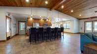 This well-appointed single-floor communal space blends rustic charm with modern functionality, making it an ideal gathering and amenity hub. The interior is unified by warm stone tile flooring, rich wood trim, and striking tongue-and-groove wood ceilings that add depth and character throughout.

At the heart of the space is a fully equipped kitchen, featuring stainless steel appliances, warm honey-toned cabinetry, light stone countertops, and a large stone-clad island with seating for several bar-height chairs. Pendant lighting above the island adds an inviting ambiance. Adjacent to the kitchen, a billiards table anchors a dedicated game area.

The lounge area offers generous seating with plush sofas and armchairs arranged around a dramatic floor-to-ceiling stacked stone fireplace with a wood mantel — a true centerpiece of the space. Decorative wall art and stylish pendant lighting complement the warm, lodge-inspired aesthetic.

A separate dining area provides high-top table seating with direct access to an outdoor patio through glass doors, allowing ample natural light to fill the room. A dedicated business center includes computer workstations, a printer, and additional table seating, framed by large windows with outdoor views.

Overall, the space exudes a welcoming, rustic-modern atmosphere well suited for socializing, working, or relaxing.
