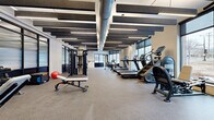 This contemporary fitness center offers a thoughtfully designed, single-floor space that accommodates a comprehensive range of workout options. The open main floor is finished with durable rubber sport flooring and features an eye-catching industrial-style ceiling with exposed ductwork, linear LED lighting, and suspended acoustic panels that serve both aesthetic and functional purposes, helping to manage sound throughout the space.

The cardio zone lines one side of the facility with a row of treadmills, stair climbers, and stationary bikes positioned to take full advantage of the large floor-to-ceiling windows, which flood the space with natural light and offer views to the outdoors. Exposed concrete columns punctuate the floor plan, adding to the facility's modern industrial character.

The strength training area is well-appointed with a cable machine system, adjustable benches, free weights, and a full rack station, providing everything needed for a complete resistance workout. A dedicated studio room with hardwood flooring, wall-mounted display, and a medicine ball storage rack offers a versatile space for functional training, stretching, or group exercises. A built-in water fountain and wood-top bench near the entrance provide added convenience for users. The combination of natural light, open layout, and high-quality equipment makes this fitness center a highly functional and inviting environment.