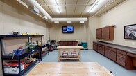 This single-floor space is thoughtfully designed to serve a variety of functional needs, combining a professional workshop, communal gathering areas, a business center, and a fully equipped kitchen under one roof.

The workshop area is a standout feature, outfitted with durable workbenches, open shelving units stocked with tools and equipment, wall-mounted storage cabinets, and a mounted display screen. Exposed ductwork and industrial fluorescent lighting run along the ceiling, complementing the utilitarian design, while a smooth sealed floor provides a clean, easy-to-maintain surface.

The lounge and common area transitions into a warm, welcoming environment, featuring rich hardwood-style flooring, warm golden-yellow accent walls, and a drop tile ceiling with recessed lighting. Large windows dressed with horizontal blinds fill the space with natural light and provide views of the surrounding landscape. Casual seating arrangements and small bistro tables create comfortable gathering spots throughout.

The business center is neatly integrated into the common area, offering workstation desks, ergonomic seating, computer monitors, and a mounted television — alongside a coffee and beverage station with dark wood cabinetry for added convenience.

The kitchen is fully functional, equipped with black appliances including a full-size refrigerator, range, and microwave, paired with a stainless steel double sink and ample cabinetry. A glazed partition connects the kitchen to an adjacent outdoor patio area, extending the usable space outdoors.