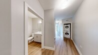 Located at 1200 Broadway Street in Ann Arbor, Michigan, this modern 2-bedroom, 2-bathroom unit offers a well-designed, single-floor layout with clean contemporary finishes throughout. The expansive living room is a true highlight, featuring floor-to-ceiling wraparound windows that flood the space with natural light and offer sweeping views of the surrounding Ann Arbor streetscape, complemented by warm hardwood-style flooring. The galley-style kitchen is thoughtfully appointed with stainless steel appliances including a French door refrigerator and gas range, two-tone cabinetry pairing white uppers with natural wood lowers, quartz countertops, an undermount sink, and a window that brings in additional natural light. Both bedrooms feature neutral tones and hardwood-style flooring; the second bedroom benefits from a large black-framed window with outdoor views. The two bathrooms are finished with white vanities, quartz countertops, large mirrors, and wood-look flooring — one bathroom includes a walk-in glass-door shower with white tile surround. A standout convenience feature is the in-unit stacked washer and dryer tucked neatly into the hallway. A dedicated closet adds practical storage to round out this well-appointed urban residence.