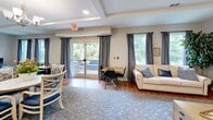 This single-floor space offers a thoughtfully designed open-concept layout that seamlessly connects the kitchen and living area, creating a welcoming and functional environment throughout. Warm hardwood flooring flows continuously across the space, complemented by neutral-toned walls and tray ceilings that add architectural interest and a sense of depth.

The kitchen is a standout feature, equipped with crisp white cabinetry, marble-style countertops, and stainless steel appliances including a large French door refrigerator and microwave. A spacious central island with bar seating for multiple guests serves as both a prep area and a casual dining hub, illuminated by a trio of pendant lights that add a modern touch.

The living area is generously proportioned and bathed in natural light from multiple large windows dressed with floor-length gray curtains. A wall-mounted flat-screen TV anchors one end of the room, while a seating arrangement featuring sofas, accent chairs, and a coffee table creates an inviting lounge atmosphere. A dedicated dining nook with a small table and chairs provides additional functionality. Sliding glass doors open to an outdoor patio area, offering pleasant views of surrounding greenery and enhancing the overall sense of openness and connectivity to the outdoors.
