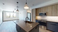 This single-floor unit presents a cohesive modern design characterized by light wood-tone flooring, crisp white walls, and bold dark accents carried consistently throughout the space. The open-concept kitchen and living area serves as the focal point, anchored by a substantial center island with a dark countertop, built-in shelving, an undermount sink, and a matte black faucet. Pendant lighting fixtures hang above the island, adding both function and visual interest. The kitchen features two-tone cabinetry pairing wood-grain upper cabinets with slate-blue lower cabinets, a white subway tile backsplash, and a full suite of stainless steel appliances including a refrigerator, range, and microwave. The bedrooms are bright and well-proportioned, each benefiting from large windows that frame outdoor views and allow generous natural light. Ceiling fans are installed in multiple rooms. The bathrooms maintain the unit's modern aesthetic, with dark vanity cabinetry, undermount sinks, dark stone-look countertops, matte black fixtures, large mirrors, and white tile surrounding the tub and shower areas. The overall ambiance is clean, contemporary, and thoughtfully detailed.