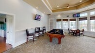 Located at 5806 Ogeechee Road in Savannah, Georgia, this single-floor community clubroom offers a sophisticated and inviting gathering space designed for both recreation and relaxation. The room's architectural character is defined by elegant tray ceilings with crown molding and warm accent detailing, recessed lighting, and classic white wainscoting that runs along the lower walls.

At the center of the space, a handsome wood-framed billiards table with a black felt surface serves as the primary recreational focal point. Along one wall, a row of high-top bar tables paired with sleek black leather stools provides additional seating, each accompanied by wall-mounted flat-screen televisions for entertainment.

A large dining area features a round wood table with upholstered arm chairs, ideal for casual gatherings or games. In a sun-drenched corner, two plush lounge chairs flank a decorative side table, positioned to take full advantage of the floor-to-ceiling windows that frame sweeping views of the outdoor pool area and surrounding greenery. Bamboo Roman shades add warmth and texture throughout.

The neutral, carpeted flooring ties the various seating zones together, creating a cohesive and comfortable atmosphere. Natural light floods the space through multiple large windows, enhancing the open, airy ambiance of this well-equipped clubroom.
