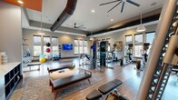 This modern fitness center offers a generously sized, open-concept layout designed to accommodate a full range of workout activities. The space is defined by soaring ceilings with exposed dark ductwork and industrial-style ceiling fans, lending the room a contemporary, loft-like aesthetic. Warm hardwood-style flooring runs throughout, complementing the neutral gray walls accented by a bold orange feature wall that adds a vibrant pop of color.

Large windows with roller shades line multiple walls, filling the space with ample natural light and providing views of the surrounding greenery. Recessed lighting and pendant fixtures ensure the space is well-lit at all hours.

The fitness floor is comprehensively equipped with a dedicated cardio zone featuring treadmills, elliptical machines, and stationary bikes. A strength training area includes a multi-station cable machine, free weight benches, and a full dumbbell rack. A functional training zone offers stability balls, medicine balls, and resistance equipment. Multiple wall-mounted flat-screen televisions are positioned throughout the space for entertainment during workouts. A built-in storage unit with open shelving provides organized equipment storage. The overall ambiance is energetic yet comfortable, striking a balance between a professional gym environment and an approachable, modern aesthetic.