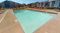The pool area at 5000 Sanger offers a generously sized rectangular swimming pool with clear, light-teal water, marked at a shallow depth of 4 feet and a slightly deeper end at 4-plus feet. The pool is finished with tiled edges and surrounded by a durable concrete sundeck, providing ample space for lounging. A row of red-orange reclining lounge chairs lines the poolside, offering residents a comfortable place to relax and soak up the sun. A wooden pergola structure anchors one corner of the deck, adding a shaded gathering spot and architectural character to the outdoor space. The pool area is enclosed by a wrought iron fence, ensuring security while maintaining open sightlines to the surrounding greenery and mature trees. The broader community grounds feature well-kept lawns, neatly trimmed hedging, and decorative rock landscaping along the building foundations. The residential buildings themselves are constructed of painted brick with a neutral palette accented by colorful exterior panel details. Individual unit entrances are accessible via concrete walkways, with metal staircases providing access between levels. The overall outdoor environment is bright and open, benefiting from abundant natural light and a spacious, park-like atmosphere.