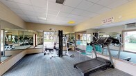 This single-floor fitness center is designed to support a well-rounded exercise routine in a functional, well-lit environment. The room is finished with a drop tile ceiling equipped with recessed panel lighting, and carpeted flooring in a neutral grid pattern provides a practical surface throughout the workout space.

The cardio section features multiple treadmills arranged in a row along one wall, all facing large wall-mounted mirrors that span the length of the room and visually expand the space. A multi-station strength training unit occupies a central position in the room, offering users options for resistance-based exercise. Additional seating is available within the space for rest periods between sets.

Large windows along two walls allow abundant natural light to enter the room while offering views of the surrounding landscaped grounds and an adjacent outdoor pool area. A keyed exterior door with full-length sidelights provides direct access to the outdoor amenities, with posted signage indicating the facility is reserved for residents only.

Wall-mounted HVAC units ensure climate control throughout the year. The mirrored walls on multiple sides of the room enhance both the sense of openness and functionality, making it easy for users to monitor their form during workouts. Overall, the space provides a practical and accessible fitness environment.