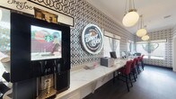 The Joe Coffee Bar at Texas A&M University is a thoughtfully designed single-floor space that seamlessly blends a café environment with a functional business center. The main café area is defined by striking black-and-white geometric wallpaper that lines the walls from floor to crown molding, creating a bold and energetic atmosphere. A long marble-topped counter runs the length of the room, lined with red upholstered bar-height chairs, offering ample seating for students and visitors. A self-service Starbucks kiosk is conveniently positioned at one end, while a glowing neon sign — "The Joe Coffee Bar, Texas A&M University" — serves as a standout focal point. Pendant globe lighting with brass accents adds warmth to the otherwise high-contrast interior. Dark hardwood flooring grounds the space throughout. An arched passageway connects the café to a dedicated business center room, which features deep navy walls, a central communal worktable with red leather chairs, and desktop computers. A large framed graphic of the London skyline adds a cosmopolitan touch to this quieter workspace. Together, the two areas create a vibrant yet practical environment suited for both casual coffee breaks and focused academic work.