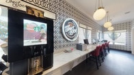 The Joe Coffee Bar at Texas A&M University is a thoughtfully designed single-floor space that seamlessly blends a café environment with a functional business center. The main café area is defined by striking black-and-white geometric wallpaper that lines the walls from floor to crown molding, creating a bold and energetic atmosphere. A long marble-topped counter runs the length of the room, lined with red upholstered bar-height chairs, offering ample seating for students and visitors. A self-service Starbucks kiosk is conveniently positioned at one end, while a glowing neon sign — "The Joe Coffee Bar, Texas A&M University" — serves as a standout focal point. Pendant globe lighting with brass accents adds warmth to the otherwise high-contrast interior. Dark hardwood flooring grounds the space throughout. An arched passageway connects the café to a dedicated business center room, which features deep navy walls, a central communal worktable with red leather chairs, and desktop computers. A large framed graphic of the London skyline adds a cosmopolitan touch to this quieter workspace. Together, the two areas create a vibrant yet practical environment suited for both casual coffee breaks and focused academic work.