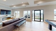 Located at 1002 Niagara St in Welland, Ontario, this thoughtfully designed single-floor open-concept space seamlessly blends a modern kitchen and living area into one cohesive, functional environment. Light hardwood flooring runs throughout, lending warmth and continuity to the space.

The kitchen is a standout feature, anchored by a substantial island topped with a striking dark stone countertop with subtle veining, surrounded by sleek navy upholstered bar stools with gold-toned legs. Custom light wood cabinetry lines the walls, complemented by a vivid blue vertical tile backsplash and arched shelving niches that add architectural character. Matte black pendant lights hang overhead, creating focused task lighting with a contemporary edge.

The living area is equally refined, centered around a built-in linear fireplace housed within a light wood media unit topped by a wall-mounted television. A custom floor-to-ceiling open shelving unit serves as both a display feature and a subtle room divider. Recessed lighting and a tray ceiling detail add dimension to the space. Large windows flood the room with natural light, while a set of black-framed glass doors opens directly onto an outdoor balcony, offering views of the surrounding area. A billiards table rounds out the recreational character of this versatile, well-appointed space.
