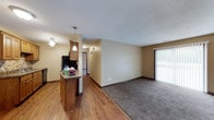 This single-floor unit is designed around an open-concept layout that seamlessly connects the kitchen and living areas, creating a cohesive and functional living space. The kitchen features medium-toned wood cabinetry, a granite-style countertop, and a peninsula that separates it from the main living area. A pendant light hangs above the peninsula, providing focused task lighting, while additional ceiling-mounted fixtures illuminate the kitchen. Black appliances add a modern contrast to the warm cabinetry tones.

The flooring transitions from hardwood-style planks in the kitchen and transitional areas to plush carpeting in the main living space, clearly delineating the two zones. Warm neutral wall tones run throughout, complemented by dark baseboards that provide a grounded visual contrast. A textured ceiling adds subtle dimension overhead.

A sliding glass door with vertical blinds fills the living area with natural light and provides access to the outdoors. A hallway visible in the background suggests additional rooms beyond the main living space. Overall, the unit offers a practical, comfortable layout with a warm and welcoming ambiance.