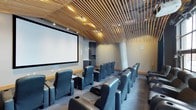 This single-floor screening room delivers a purpose-built cinematic environment with thoughtful design throughout. The most striking architectural feature is the sweeping slatted wood ceiling, where angled timber slats cascade dramatically toward the front of the room, providing both aesthetic warmth and acoustic functionality. Recessed lighting integrated between the slats casts a warm, ambient glow across the space.

Rows of plush, high-back leather recliner seats in a deep blue-grey tone are arranged in a tiered layout facing a large, wall-mounted projection screen. Each seat features integrated armrests with cup holders, prioritizing comfort for extended viewing sessions. The tiered arrangement ensures unobstructed sightlines from every position in the room.

The neutral grey carpeted flooring and soft grey walls create a clean, distraction-free backdrop that enhances the viewing experience. At the rear of the room, a compact built-in cabinetry unit with a microwave and storage provides convenient refreshment access. Subtle wall-mounted speakers support the room's audio setup. Overall, the space combines modern design with functional amenities to create a polished, communal screening experience.