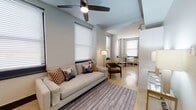 This well-designed single-floor space offers a thoughtful layout that flows seamlessly from room to room. The living area is bright and welcoming, featuring large windows with white blinds that invite natural light throughout the day. A ceiling fan overhead adds both style and function, while polished stone-look flooring ties the space together. The living area is furnished with a generous neutral sofa accented by patterned throw pillows, a mid-century style armchair in warm brown, and a classic floor lamp, creating an inviting and relaxed atmosphere. Adjacent to the living area, a dedicated dining nook with a round table and chairs provides an intimate space for meals.

The kitchen is clean and contemporary, outfitted with white cabinetry, light quartz countertops, a white tile backsplash, and a full suite of stainless steel appliances including a range, dishwasher, and microwave. The bedroom offers a calm retreat with neutral gray walls, dark wood flooring, and a platform bed framed by a modern white paneled headboard. The bathroom is finished with wood-look tile flooring, a white vanity, and a walk-in shower enclosed by frameless glass panels with white subway tile surround — a clean, minimalist design throughout.