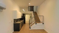 This well-designed two-story space combines modern aesthetics with functional living across two thoughtfully laid-out floors. The main level showcases an open-concept kitchen that serves as a visual centerpiece, outfitted with dark lower cabinetry contrasted by crisp white upper cabinets. A dark tile backsplash adds texture and depth, while stainless steel appliances — including a refrigerator, dishwasher, and built-in microwave — lend a polished, contemporary feel. The countertops offer ample workspace, complemented by a stainless steel sink beneath recessed and fluorescent lighting that keeps the area bright and practical. Warm-toned hardwood-style flooring flows throughout the main level, adding a natural warmth that balances the space's modern palette of black, white, and grey. A standout architectural feature is the open-riser staircase with rich wood treads and sleek metal railings, rising dramatically toward the upper floor and drawing the eye upward to the impressively tall ceilings above. The generous ceiling height amplifies the sense of openness and airiness throughout. A hallway off the main area leads to additional spaces on the ground floor, while the staircase provides access to the second level, completing a versatile and stylish multi-floor layout.