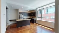 This contemporary single-floor residence offers a well-designed open-concept layout that seamlessly blends functionality with modern style. The kitchen serves as the centerpiece of the living space, showcasing rich dark espresso cabinetry that extends to the ceiling for ample storage, granite countertops with a light speckled finish, and a full suite of stainless steel appliances including a refrigerator, electric range, and over-the-range microwave. A peninsula with a built-in sink and matching granite surface provides additional prep space and doubles as a casual dining or entertaining area. A white subway tile backsplash adds a clean, polished accent to the kitchen walls. Warm hardwood flooring flows throughout the main living areas, reflecting abundant natural light that pours in through large floor-to-ceiling windows overlooking an urban streetscape. The bathroom is finished with consistent attention to detail, featuring a dark wood vanity topped with granite, a framed mirror, tile flooring, and a full soaking tub with a tiled surround and curved shower rod. A dedicated in-unit laundry closet houses a stacked Bosch washer and dryer, offering a premium and convenient amenity. Recessed lighting throughout completes the clean, contemporary aesthetic of this thoughtfully appointed space.