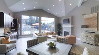 This single-floor space features a dramatic vaulted ceiling with recessed lighting that creates an airy, open atmosphere throughout. Large floor-to-ceiling windows along one wall flood the interior with natural light while offering expansive views of an outdoor pool area, complete with lounge seating and mature trees beyond a perimeter fence.

The main living area is anchored by a sleek, tile-surround fireplace topped with a wall-mounted flat-screen television, serving as a refined focal point. Neutral gray walls complement the light-toned flooring and contribute to the space's calm, contemporary aesthetic.

Comfortable seating arrangements — including sofas, armchairs with patterned upholstery, and side tables — are thoughtfully positioned around a central area rug, encouraging conversation and relaxation. A large potted plant adds a touch of greenery and warmth to the room.

A glass-topped dining table with upholstered chairs anchors a defined dining zone in the foreground, complete with a decorative centerpiece bowl. The overall design balances functionality with an inviting, polished ambiance suited for both casual gatherings and quiet retreats.