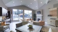 This single-floor space features a dramatic vaulted ceiling with recessed lighting that creates an airy, open atmosphere throughout. Large floor-to-ceiling windows along one wall flood the interior with natural light while offering expansive views of an outdoor pool area, complete with lounge seating and mature trees beyond a perimeter fence.

The main living area is anchored by a sleek, tile-surround fireplace topped with a wall-mounted flat-screen television, serving as a refined focal point. Neutral gray walls complement the light-toned flooring and contribute to the space's calm, contemporary aesthetic.

Comfortable seating arrangements — including sofas, armchairs with patterned upholstery, and side tables — are thoughtfully positioned around a central area rug, encouraging conversation and relaxation. A large potted plant adds a touch of greenery and warmth to the room.

A glass-topped dining table with upholstered chairs anchors a defined dining zone in the foreground, complete with a decorative centerpiece bowl. The overall design balances functionality with an inviting, polished ambiance suited for both casual gatherings and quiet retreats.