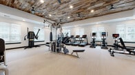 This fitness center is a thoughtfully designed, single-floor facility that blends rustic aesthetic charm with modern functionality. The standout architectural feature is the expansive reclaimed wood plank ceiling, which adds warmth and character throughout the main workout areas, complemented by recessed lighting and ceiling fans for optimal comfort. The space is divided into several well-defined workout zones, each catering to different fitness disciplines. The main floor houses a generous array of cardio equipment, including multiple treadmills, ellipticals, stair climbers, and stationary bikes, all positioned near large windows with blinds that allow in ample natural light. A dedicated strength training area features a Hoist multi-station cable machine, weight benches, a full dumbbell rack, and resistance equipment. A functional training zone is equipped with a punching bag, battle ropes, TRX suspension trainers, plyometric boxes, a squat rack with accessories, and a rowing machine. A separate, enclosed room with glass-panel doors offers additional specialty equipment, including a cable tower machine and built-in cubby storage. The facility also includes a corridor entry with glass-framed doors, a living green wall accent, and direct outdoor access through French doors, creating a bright, airy atmosphere throughout.