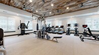 This fitness center is a thoughtfully designed, single-floor facility that blends rustic aesthetic charm with modern functionality. The standout architectural feature is the expansive reclaimed wood plank ceiling, which adds warmth and character throughout the main workout areas, complemented by recessed lighting and ceiling fans for optimal comfort. The space is divided into several well-defined workout zones, each catering to different fitness disciplines. The main floor houses a generous array of cardio equipment, including multiple treadmills, ellipticals, stair climbers, and stationary bikes, all positioned near large windows with blinds that allow in ample natural light. A dedicated strength training area features a Hoist multi-station cable machine, weight benches, a full dumbbell rack, and resistance equipment. A functional training zone is equipped with a punching bag, battle ropes, TRX suspension trainers, plyometric boxes, a squat rack with accessories, and a rowing machine. A separate, enclosed room with glass-panel doors offers additional specialty equipment, including a cable tower machine and built-in cubby storage. The facility also includes a corridor entry with glass-framed doors, a living green wall accent, and direct outdoor access through French doors, creating a bright, airy atmosphere throughout.