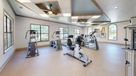 Located in Cordova, TN, this 24-hour fitness facility offers a thoughtfully designed workout environment on a single floor. The main exercise area is generously proportioned, providing ample room to accommodate a diverse array of fitness equipment, including treadmills, elliptical trainers, stationary bikes, and a cable strength-training machine with a dedicated bench. The space is brightened by multiple large windows, including a distinctive arched window, which fill the room with natural light and offer views of the surrounding greenery. The ceiling features an eye-catching coffered design accented in blue and wood tones, with dual ceiling fans ensuring comfortable airflow throughout workouts. Recessed lighting and crown molding add refined architectural detail to the space. Wall-mounted televisions and framed artwork contribute to an engaging atmosphere. Light-toned flooring runs throughout, complementing the clean, neutral walls. A dedicated bathroom provides added convenience for users. A glass-panel exterior door offers direct outdoor access, making entry and exit effortless. Overall, the facility blends functional fitness amenities with polished architectural finishes, creating an inviting and well-appointed environment for round-the-clock exercise.