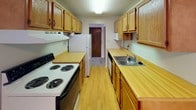 This single-floor unit at 9727 Pleasant Lake Blvd in Parma, Ohio, offers a straightforward, functional layout across several distinct areas. The galley-style kitchen is well-equipped with warm oak cabinetry lining both walls, light wood laminate flooring, yellow-toned countertops, a double-coil electric range, a built-in microwave, a dishwasher, and a stainless steel sink. Ceiling fans are present throughout the unit, providing consistent air circulation. The living area features neutral carpet, large windows dressed with vertical blinds, and baseboard heating, allowing ample natural light to filter in. An open layout connects the kitchen to a secondary living or dining space, also finished with light hardwood-style flooring and a ceiling fan. The bedroom areas are carpeted with textured ceilings, ceiling fans, and windows with vertical blinds. Closet access is provided via wood-paneled sliding doors. The bathroom includes a vanity with a wide countertop, a large wall-spanning mirror with a lit chrome fixture above, tile flooring, and a tub-shower combination. Overall, the unit presents a clean, utilitarian aesthetic suited for comfortable everyday living.