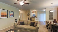 This single-story space showcases an open-concept layout that seamlessly connects the living room, dining area, and kitchen. The living room serves as the central gathering space, highlighted by crown molding along the ceiling perimeter and a ceiling fan with a light fixture that provides both style and air circulation. Neutral beige walls and warm-toned flooring create a cohesive, inviting atmosphere throughout.

The living area is generously proportioned, offering ample room for comfortable seating arrangements. Natural light filters in through a large set of vertical blinds along one wall, brightening the space considerably. A smooth transition leads into the dining area, where a dark-topped breakfast bar provides casual seating with bar-height chairs, creating a natural division between the living and kitchen spaces.

The kitchen is visible from the main living area and features white cabinetry, a dark appliance such as a built-in microwave or oven, and recessed track lighting overhead. The open sightlines between rooms enhance the sense of spaciousness and allow for easy interaction across the living, dining, and kitchen zones, making the layout both functional and socially connected.