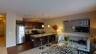 This single-floor space showcases an open-concept layout that seamlessly integrates the kitchen, dining, and living areas into one cohesive environment. The kitchen is outfitted with rich dark-wood cabinetry, granite countertops, and stainless steel appliances, including a full-size refrigerator, built-in microwave, and range. A central island with bar-height seating serves as both a functional prep space and an informal dining area, illuminated by a pair of elegant pendant lights that hang overhead.

Track lighting along the kitchen ceiling provides additional task lighting, while warm ambient lighting throughout creates an inviting atmosphere. The transition from kitchen to living area is fluid, anchored by a bold zebra-print area rug that adds a dramatic design element. The living space features a tufted sofa with neutral upholstery, a glass coffee table, and a large decorative mirror that enhances the sense of depth. Warm-toned walls and hardwood flooring unify the space, contributing to an overall ambiance that is both modern and elegantly appointed.