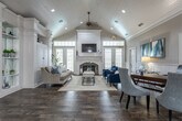 Located in Gainesville, Georgia, this single-story community space combines classic Southern architecture with refined coastal-inspired design. The welcoming entryway greets visitors with rich hardwood flooring, white decorative columns, crown molding, and a striking pendant chandelier that sets the tone for the interior. Large glass-paneled front doors with sidelights flood the foyer with natural light, while navy blue drapery adds a bold, polished accent.

The main living area is a standout, featuring soaring vaulted ceilings finished with white beadboard paneling and recessed lighting. A central fireplace with an elegant white surround and dark stone hearth serves as the focal point of the room, complemented by a wall-mounted television above the mantle. Comfortable seating, including a neutral linen sofa and deep navy accent chairs, creates an inviting gathering space framed by multiple French doors and windows.

A private office area offers a professional setting with a classic wood desk, upholstered guest chairs, and glass-paned interior doors that maintain an open, airy feel while providing privacy. Throughout the space, consistent use of white wainscoting, soft gray walls, and dark hardwood floors creates a cohesive, sophisticated aesthetic. The overall ambiance is bright, elegant, and welcoming.