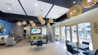 This single-floor communal space is designed with a contemporary aesthetic that balances style and functionality. The ceiling is a standout architectural feature, showcasing a coffered design finished in a bold dark tone, which contrasts sharply with the lighter walls and flooring below. Suspended throughout the space is a dramatic collection of ornate gold spherical pendant lights, varying in size and adding a warm, sculptural element that draws the eye upward and anchors the room's modern design language.

The space is organized into distinct zones that serve different purposes. A dining area features sleek tables paired with charcoal and lime-green chairs, creating a vibrant, energetic corner ideal for casual gatherings. Adjacent to this is a large wall-mounted flat-screen television set against a textured stone accent wall, flanked by what appears to be a kitchenette or bar area with stainless steel appliances.

Toward the exterior wall, a comfortable lounge seating area with neutral-toned armchairs and sofas encourages relaxed conversation. Floor-to-ceiling glass doors flood this zone with natural light and offer views of an outdoor area, creating a seamless indoor-outdoor connection. The overall ambiance is polished, welcoming, and well-suited for social interaction.