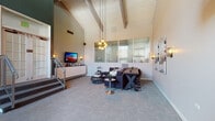 This two-story amenity facility is defined by its striking architectural character, most notably the soaring vaulted ceiling with exposed white-painted wood beams that draws the eye upward throughout the main lounge area. The ground floor opens into a spacious common lounge furnished with plush seating, mixed accent tables, and a wall-mounted television, creating a welcoming gathering space. Pendant cluster lighting descends dramatically from the peak of the vault, adding warmth alongside wall sconces. A row of large frosted-glass transom windows along an interior wall allows diffused light to pass between areas while maintaining visual separation. Near the main entrance, a dining area with a long table and chairs sits beside tall windows overlooking the outdoors. A well-appointed kitchen features rich dark cabinetry, black granite countertops, a metallic tile backsplash, and stainless steel appliances. A dedicated fitness center offers a range of strength and cardio equipment, wall mirrors, and ample floor space. A staircase with metal railings leads to the upper level, where a loft-style co-working nook provides desk stations beneath the sloped ceiling, decorated with vibrant artwork—an ideal space for focused work above the activity below.