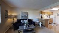 This single-floor residence offers a well-designed, open-concept layout that promotes a sense of connectivity and ease of movement throughout the space. The living area serves as the heart of the home, featuring neutral-toned walls and carpeted flooring that create a warm and inviting atmosphere. A textured ceiling adds subtle architectural character overhead. The space comfortably accommodates a seating arrangement centered around a modern coffee table, making it well-suited for both relaxing and entertaining. A decorative cityscape wall print adds a contemporary artistic touch to the living area. The kitchen is seamlessly integrated into the open floor plan via a raised breakfast bar with counter seating, fostering a sociable connection between the cooking and living spaces. The kitchen features warm wood-toned cabinetry, a light-colored countertop, and essential appliances including a built-in microwave. The overall ambiance of the space is casual and functional, with a neutral color palette throughout that offers a versatile backdrop for personalizing the interior. The single-floor layout ensures convenient, step-free access to all areas of the home.