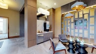 This elegantly designed single-floor space serves as a welcoming lobby and lounge area, combining contemporary aesthetics with functional amenities. The interior is anchored by warm wood-paneled columns and cabinetry that frame a built-in refreshment station, complete with a marble-topped counter, beverage equipment, and under-counter refrigeration — all finished to a high standard. Recessed lighting and statement pendant fixtures with gold-toned interiors cast a warm, inviting glow throughout the space.

The lounge area is thoughtfully appointed with a curated mix of upholstered seating, including tufted accent chairs in velvet and fabric, arranged around low tables to encourage relaxed conversation. A decorative lattice-style partition adds visual depth and architectural interest to the room's layout. An adjacent seating nook is bathed in natural light from a dramatic floor-to-ceiling arched window wall, offering views of a lush green landscape and streetscape beyond. Sheer draping curtains flank the windows, softening the light while maintaining an open, airy feel.

Contemporary abstract artwork is displayed prominently on the walls, adding bold splashes of color against the neutral palette of soft beige walls and large-format tile flooring. Textured stone accent walls provide additional contrast and tactile richness. Together, these elements create a polished, welcoming environment with a refined yet approachable character.