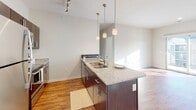 This single-floor unit showcases a contemporary design with a cohesive, neutral color palette throughout. The open-concept kitchen serves as a central highlight, featuring a large island with granite countertops and an integrated sink, dark espresso cabinetry, and a full suite of stainless steel appliances, including a refrigerator, electric range, and microwave. Pendant lighting hangs above the island, while a track lighting system provides additional illumination. The kitchen flows seamlessly into the living area, which benefits from abundant natural light and warm wood-look flooring that extends throughout the main living spaces.

The bedroom offers a quieter atmosphere, finished with plush neutral carpeting and a window with blinds that allows for controlled natural light. The room's clean walls and open layout provide a versatile canvas for furnishing.

The bathroom is well-appointed with dark espresso vanity cabinetry, a granite-topped sink, a large mirror, and a full tub-shower combination with a curtain rod. Wood-look flooring continues into this space, maintaining design consistency. The overall layout, as seen in the floor plan view, is efficiently organized, connecting the kitchen, living area, bedroom, and bathroom in a practical and accessible arrangement.