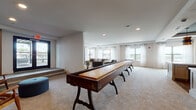 This well-appointed single-floor amenity space is designed for socializing and recreation, offering a diverse range of zones that cater to both casual gatherings and lively entertainment. The open layout flows seamlessly from one area to the next, all finished with neutral tones, recessed lighting, and plush carpeting throughout most of the space.

The game room is a standout feature, centered around a full-length wooden shuffleboard table. Multiple large windows dressed with sheer white curtains fill the room with natural light and offer views of the surrounding cityscape. Adjacent to the game room is a comfortable lounge area furnished with a neutral sofa, mid-century modern accent chairs, and a round coffee table — all oriented toward a large wall-mounted television framed by expansive floor-to-ceiling windows.

The catering kitchen is both functional and elegant, featuring white shaker cabinets, white quartz countertops, and rich hardwood flooring. A large central island is illuminated by a striking cluster of gold and black pendant lights. Wall-mounted televisions and a mirrored backsplash add modern flair, while ample counter seating and a wet bar make it ideal for hosting events.

A dedicated bar counter area lined with dark wood barstools sits beneath a graphic Atlanta city map print, adding a local touch to the design. The space combines practicality with refined aesthetics throughout.