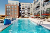 Located at 3305 South Blvd in Charlotte, NC, this dynamic outdoor amenity space is the centerpiece of a modern, multi-story residential community. The architecture blends classic brick facades with contemporary gray paneling and dark metal accents, creating an urban yet refined aesthetic that frames the open-air courtyard on all sides.

At the heart of the space is a generously sized resort-style swimming pool with crystal-clear blue water, accessible via multiple entry points and flanked by a broad, light-toned concrete sundeck. The deck is well-appointed with rows of white and charcoal lounge chairs, vibrant red umbrellas for shade, and private canopy cabanas, offering a variety of relaxation options under the open sky.

A separate outdoor terrace area showcases curated landscaping with ornamental grasses, low shrubs, and raised planting beds lining a wide paved walkway. A pergola-style architectural feature anchors this space, accompanied by lounge seating and an outdoor entertainment screen, making it ideal for both quiet relaxation and social gatherings.

The surrounding multi-story building features private balconies on upper floors, providing elevated views of the courtyard below. The overall ambiance is bright, open, and resort-inspired, with abundant natural light enhancing the inviting atmosphere of this well-designed outdoor retreat in the heart of Charlotte.