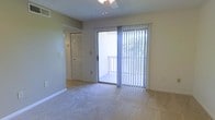 This single-story space features a bright and simply appointed living area that serves as the heart of the layout. The room is finished with wall-to-wall carpeting that extends toward a tiled transition area near the sliding glass door, which opens directly onto an outdoor balcony enclosed by a white railing. Vertical blinds frame the sliding door, offering privacy while allowing natural light to filter into the room. A ceiling fan is mounted overhead, providing both lighting and airflow throughout the space. The textured ceiling adds a subtle design element consistent with classic residential construction. Neutral green-toned walls keep the space feeling calm and understated. Adjacent to the main living area, a hallway leads further into the unit, where a louvered bi-fold closet door is visible, suggesting convenient storage access. The overall layout is open and functional, with a clean, move-in ready presentation. The connection to the outdoor balcony enhances the sense of space and brings in views of surrounding greenery, adding a refreshing natural element to the interior ambiance.