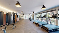 Located at 1105 Pleasant St in Oak Park, IL, this modern fitness center offers a thoughtfully designed, single-floor layout spanning a generously sized open floor plan. The main workout area features large-format rubber flooring tiles and is lined with an impressive array of TRUE-brand cardio equipment, including treadmills, ellipticals, and stair climbers positioned along floor-to-ceiling windows that flood the space with natural light and offer views of the surrounding neighborhood. The strength training section is well-appointed with a power rack, cable machines, free weights, and adjustable benches, all arranged to allow ample room for movement. Warm-toned accent walls in terracotta hues and full-length mirrors add depth and visual interest throughout the space. Industrial-style pendant lighting provides consistent, even illumination across the entire floor. A separate studio room features plush carpet flooring, additional large windows, a spin bike, a mini trampoline, and a rack stocked with kettlebells, resistance equipment, and yoga accessories — ideal for group classes or individual training. A frosted glass partition elegantly divides the studio from the main gym. The overall ambiance is clean, bright, and professional, reflecting a well-maintained facility designed to support a full range of fitness activities.