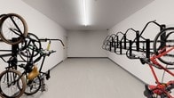 This dedicated bike storage room is a well-organized, single-floor amenity space designed to accommodate multiple bicycles in an efficient and secure manner. The room features wall-mounted vertical bike racks lining both sides, allowing bicycles to be stored upright and maximizing the use of available floor and wall space. The racks are constructed of sturdy black metal and are capable of holding a variety of bicycle types and sizes.

The interior is finished with smooth, light-colored walls and a clean light gray floor, giving the space a bright and tidy appearance. Overhead, linear fluorescent lighting runs the length of the ceiling, ensuring the room is well-lit throughout. The ceiling height provides comfortable clearance for handling and mounting bikes on the racks.

Access to the room is provided through a solid, secure door equipped with a door closer mechanism, ensuring the space remains closed and protected at all times. A security camera is also visible, adding an additional layer of safety for stored bicycles.

The overall design of the space prioritizes functionality and practicality, making it a convenient amenity for cyclists. The clean, minimalist aesthetic keeps the focus on usability, while the organized layout ensures that multiple bikes can be stored without clutter, making it easy to retrieve and return bicycles as needed.