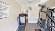 This fitness center is a well-appointed, single-level exercise space designed to support a range of workout routines. The room is finished with warm beige walls, crown molding, and carpeted flooring, creating a clean and comfortable environment. Natural light filters in through multiple large windows fitted with horizontal blinds, as well as through glass-paned French doors that provide direct access to the outdoors, offering views of a green exterior landscape.

The space is equipped with a multi-station Apollo strength training machine, which anchors the room and accommodates a variety of resistance exercises. A TrueForm treadmill is positioned along one wall, providing dedicated cardio options, while a stationary air bike adds an additional aerobic training option. A wall-mounted monitor is installed near the treadmill area for convenience.

Overhead, flush-mounted lighting supplements the natural light, ensuring the room remains bright and functional at all times. A posted fitness center rules placard indicates that this is a shared or community-accessible facility. The room connects to adjacent interior spaces through a standard doorway, and the overall layout is efficient and practical for focused physical training.