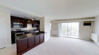 This single-floor space offers a thoughtfully designed open-concept layout that seamlessly connects the kitchen and living areas. The kitchen is outfitted with rich dark wood cabinetry, dark granite countertops, and stainless steel appliances, including a built-in microwave and dishwasher. Track lighting above the kitchen provides focused illumination, while a breakfast bar peninsula offers additional counter space and serves as a natural divider between the kitchen and the main living area. The living area features light neutral carpeting that adds warmth and comfort underfoot. Large floor-to-ceiling sliding glass doors dominate the far wall, flooding the space with abundant natural light and providing direct access to an outdoor balcony. The views through the glass doors reveal a lush, tree-lined outdoor setting, enhancing the sense of openness and connection to the outdoors. The overall color palette is clean and neutral, with white walls and ceilings that complement the contrast of the dark kitchen finishes. The open floor plan and generous proportions create a versatile, airy atmosphere well-suited for both everyday living and entertaining.