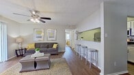 This single-story residence offers a thoughtfully designed layout centered around an open-concept living and kitchen area. The living room features warm wood-style flooring, a ceiling fan with integrated lighting, and a textured ceiling. A large sliding glass door floods the space with natural light and provides access to an outdoor area. The kitchen is a galley-style design outfitted with granite countertops, white cabinetry, stainless steel appliances including a refrigerator, microwave, and dishwasher, and a decorative tile backsplash. A pass-through counter opens to the living area and is complemented by a row of bar-height stools, creating a casual dining option. A separate dining nook accommodates a round table and chairs alongside a window. The bedrooms are carpeted and each includes a ceiling fan, natural light from windows, and closet access. The bathrooms are equipped with granite-topped vanities and a tub-shower combination. Throughout the residence, a consistent neutral gray color palette, coordinated accents, and wood-style flooring in main areas create a cohesive and inviting ambiance.