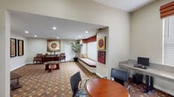 This single-floor amenity space is thoughtfully designed to serve as a multifunctional community hub, blending comfort, productivity, and wellness in one cohesive environment. The interior is anchored by a distinctive geometric-patterned carpet in warm earth tones — rich browns, reds, and golds — that flows throughout the entire space, creating a unified and visually engaging aesthetic.

The lounge area offers a welcoming atmosphere with a circular wood coffee table, leather accent chairs, and a console table adorned with a bold circular abstract artwork. A tall potted palm adds a touch of greenery, while recessed lighting illuminates the neutral beige walls. A built-in window bench with a cushioned seat and Roman shade provides a cozy nook for relaxing.

Adjacent to the lounge is a dedicated business center featuring a long wall-mounted desk with computer workstations, complemented by a round meeting table surrounded by modern rolling chairs. Colorful abstract artwork and windows with natural light enhance the work-friendly environment.

A wall-mounted flat-screen television and framed artwork occupy another seating area, offering a casual media space. Large interior windows look directly into a well-equipped fitness center, which is accessible through a private door and features treadmills and various exercise machines, making wellness both visible and convenient.