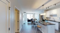 This contemporary single-floor residence showcases a thoughtfully designed open-concept layout that effortlessly connects the kitchen, dining, and living spaces into one cohesive environment. The interior is finished with sleek hardwood-style flooring that runs throughout, complementing the crisp white walls and clean architectural lines that define the space.

The kitchen features a dark-topped breakfast bar with seating for two, white cabinetry, and modern pendant lighting overhead — ideal for casual dining or entertaining. Adjacent to the kitchen, a compact dining area accommodates a round table with stylish chairs, creating a natural transition point between cooking and relaxing.

The living area anchors the far end of the space, bathed in natural light streaming through large windows that offer views to the outside. The layout is open and airy, with ample room for comfortable seating arrangements. A soft, neutral color palette is carried throughout, accented by modern furnishings in deep navy and warm tan tones.

A hallway near the entrance provides access to additional rooms, with clean door frames and subtle architectural details that maintain the modern aesthetic. Overall, the space presents a bright, functional, and stylish environment suited for contemporary everyday living.