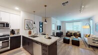 This well-appointed single-floor residence showcases a thoughtfully designed open-concept layout that seamlessly connects the kitchen, dining, and living areas. The kitchen is a standout feature, boasting a large granite-topped island with an undermount sink and seating for two, complemented by sleek dark cabinetry on the lower level and crisp white upper cabinets. A full suite of stainless steel appliances — including a French door refrigerator, gas range, built-in microwave, and dishwasher — adds a polished, functional edge. Stylish pendant lighting with matte black fixtures illuminates the island and adds a modern touch. Light wood-look flooring flows throughout the main living areas, creating a warm, cohesive aesthetic. The living room offers generous space with large windows that invite abundant natural light and feature neutral-toned walls that enhance the bright, airy ambiance. Two well-sized bedrooms each include ceiling fans, plush carpeting, and windows with blinds and sheer curtains. Both bathrooms mirror the home's refined design language, featuring granite vanity countertops, dark-framed mirrors, undermount sinks, and walk-in glass-enclosed showers, providing a clean and contemporary finish throughout.